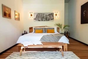 A modern bedroom with a large bed featuring white bedding, striped and yellow pillows, a wooden bench at the foot, wall-mounted nightstands, art on the walls, and a patterned rug on a wooden floor.