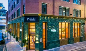 A modern corner building with green tile facade and large windows glows warmly at dusk. A black sign above the door reads Wilde. The street is quiet with minimal traffic and city buildings nearby.