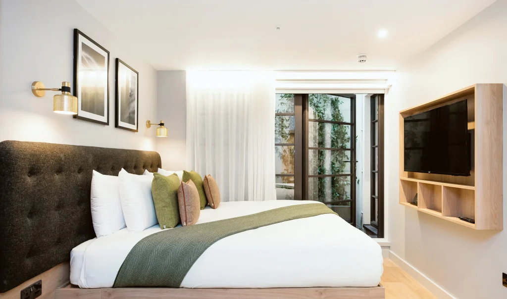 Modern hotel room with a large bed, green throw and pillows, wall-mounted lamps, framed art, a flat-screen TV, and glass doors leading to a patio with greenery outside. The room has bright, natural lighting.