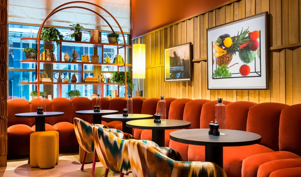 A stylish café interior with curved red velvet seating, round black tables, patterned chairs, and decorative shelving with plants and books. Framed artwork and warm lighting create a cozy, modern atmosphere.