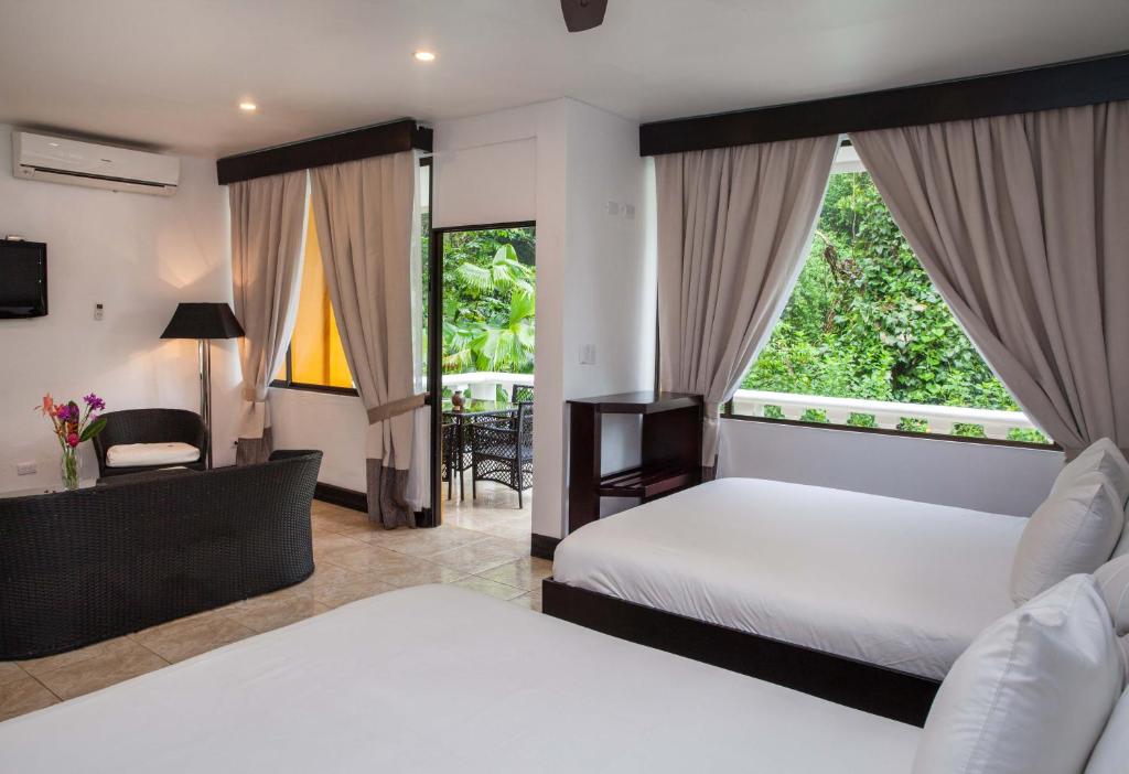 A bright hotel room with two white beds, a wicker sofa, a dark wood shelf, large windows with beige curtains, and a door opening to a leafy outdoor patio.