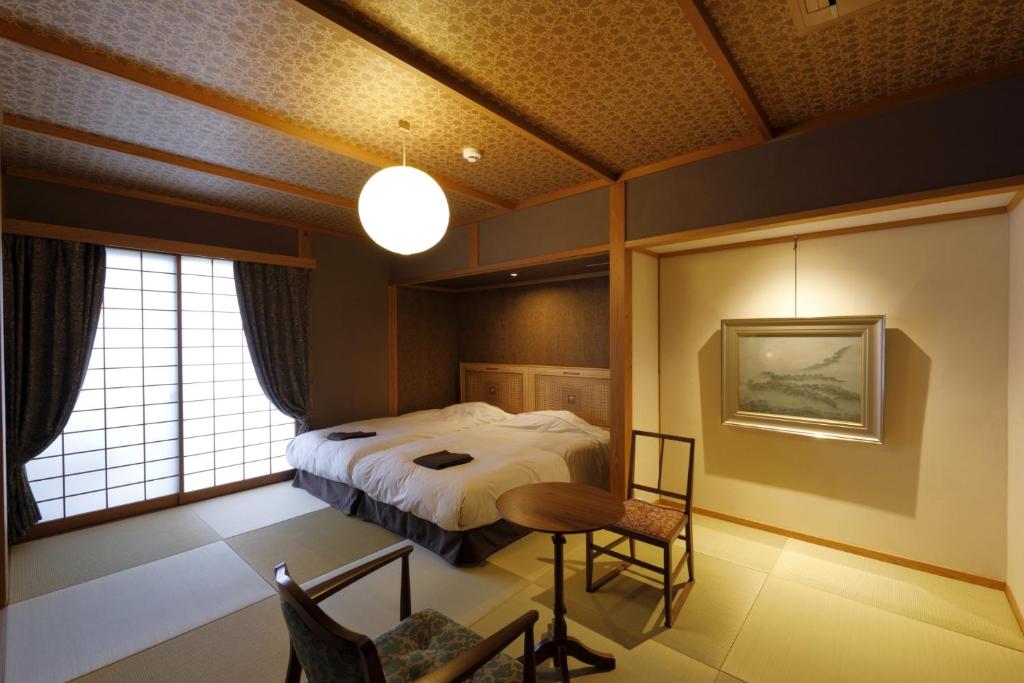 A traditional Japanese-style room with tatami flooring, two twin beds, a round wooden table with two chairs, shoji screen doors, and a framed artwork on the wall. Soft lighting creates a warm atmosphere.