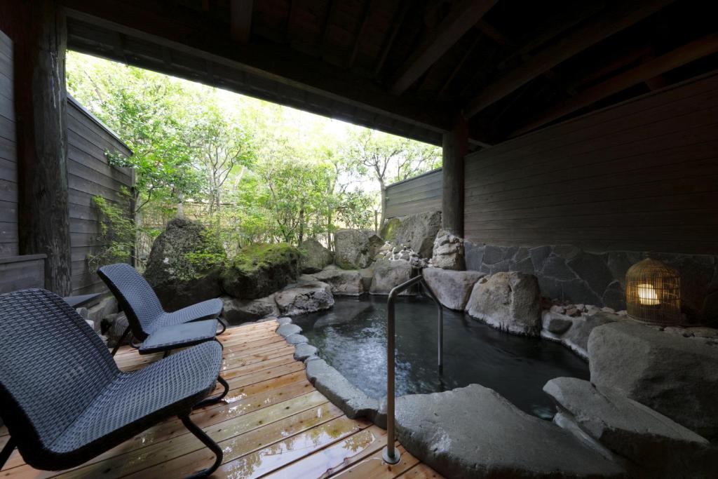 Outdoor hot spring bath with large rocks and natural wooden decking, three black wicker chairs, and greenery visible through an open wall under a wooden roof. A lantern sits on a stone ledge by the water.