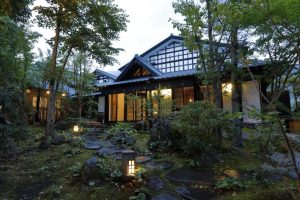 A traditional Japanese house with wooden beams and large windows is surrounded by lush greenery and stone lanterns along a mossy garden path at dusk.