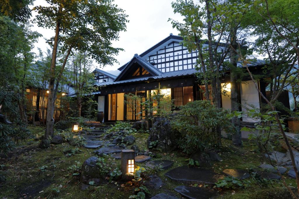 A traditional Japanese house with wooden beams and large windows is surrounded by lush greenery and stone lanterns along a mossy garden path at dusk.