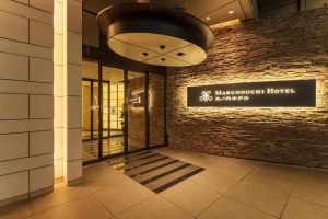 Entrance to Marunouchi Hotel with warm lighting, stone wall, large illuminated sign, and glass doors leading inside. The area has a modern, upscale appearance with clean lines and elegant design.