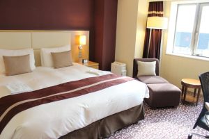 A neatly made hotel room features a large bed with white linens and brown accents, two pillows, a lamp on the wall, a small nightstand, an armchair with an ottoman, and a window letting in natural light.