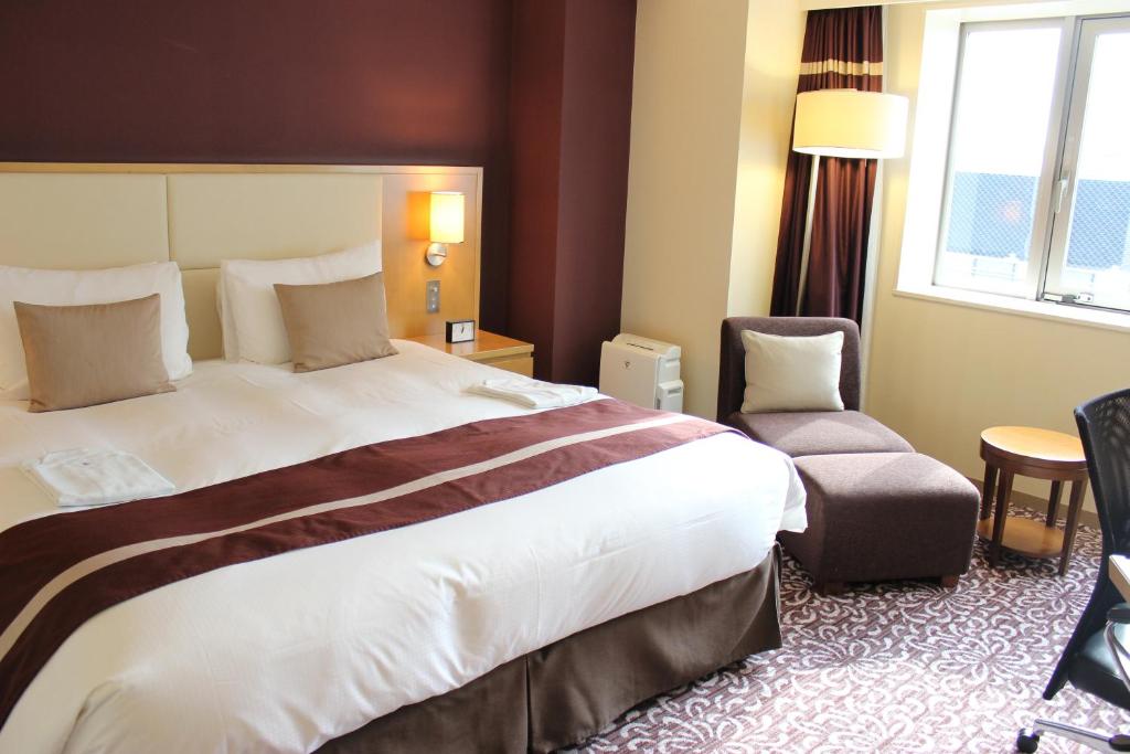 A neatly made hotel room features a large bed with white linens and brown accents, two pillows, a lamp on the wall, a small nightstand, an armchair with an ottoman, and a window letting in natural light.