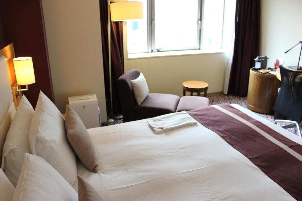 A neatly made hotel bed with white linens and brown accents, a bedside lamp, an armchair with a small round table by the window, and a desk with a chair and coffee maker in the corner.
