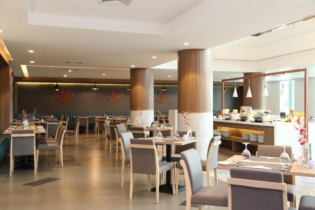 A modern, well-lit restaurant dining area with wooden tables, grey chairs, and columns. Place settings are neatly arranged, and a buffet area with food and plates is visible in the background. Subtle wall art decorates the space.
