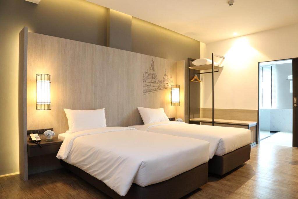 Modern hotel room with two neatly made single beds, light wood paneling, wall lamps, open closet space, wooden floor, and a view into a bright bathroom area. The room has warm, ambient lighting.