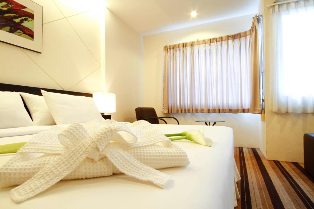 A neatly made bed with white linens and a folded robe is in a bright hotel room. There is a chair, small table near the window with beige curtains, and a painting on the wall above the bed.