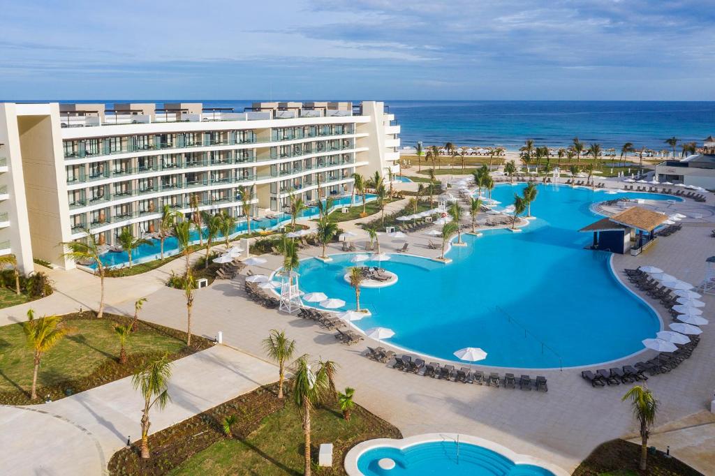 A modern beachfront resort features a large, curving swimming pool surrounded by lounge chairs, palm trees, and a multi-story hotel building. The ocean and sandy beach are visible in the background under a partly cloudy sky.