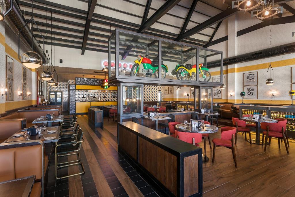 Modern restaurant interior with wooden floors, red upholstered chairs, brown booths, and tables set for dining. A glass display above features two colorful motorcycles. Industrial ceiling and decorative lighting are visible.