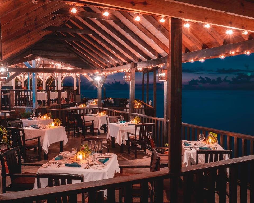 A romantic outdoor restaurant with wooden beams, string lights, and white-tableclothed tables set for dinner, overlooking a calm ocean at dusk. Warm candlelight creates a cozy, inviting ambiance.