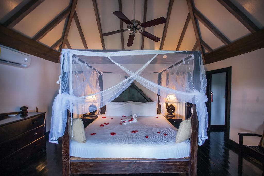 A cozy bedroom with a high wooden ceiling and a four-poster bed draped in white mosquito netting. The bed is decorated with rose petals and towel art. Two lamps sit on bedside tables, and a ceiling fan hangs above.