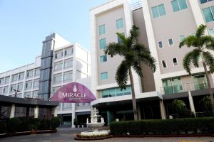 A modern multi-story hotel building with palm trees in front, a central white fountain, and a purple sign reading Miracle Airport Hotel. The exterior is light-colored with many windows.