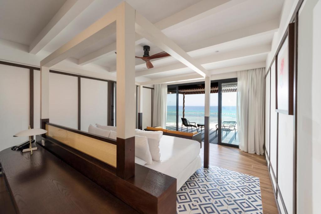A modern, airy bedroom with a four-poster bed, wooden floors, and a geometric rug. Large glass doors open to a balcony with ocean views, lounge chairs, and natural light streaming in.