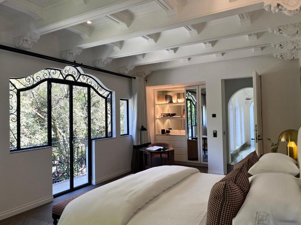A bright, elegant bedroom features a large bed, an ornate ceiling, floor-to-ceiling windows with wrought-iron details, a small desk, built-in shelves, and a view into a hallway with arched doorways.