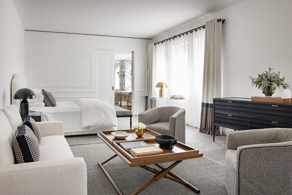 A modern, minimalist bedroom with white walls, a white bed, neutral-tone furniture, a wooden coffee table, and large windows with sheer curtains, creating a bright and airy atmosphere.