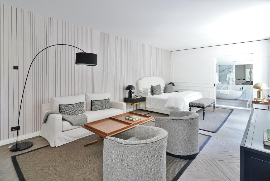 A modern hotel room with a white bed, a white sofa, two gray armchairs, a wooden coffee table, a floor lamp, and an open bathroom with a bathtub in the background. The decor is minimalist and neutral-toned.