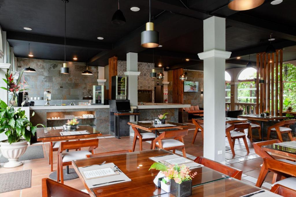 A modern, well-lit restaurant with wooden tables and chairs, glass tabletops, potted plants, and large windows letting in natural light. The counter area has coffee machines and decorations.