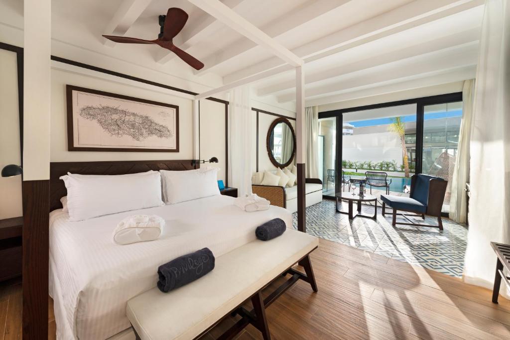 Bright hotel room with a canopy bed, white bedding, towels, a bench, ceiling fan, wall art, sofa, round mirror, balcony with chairs, and large windows letting in natural light.