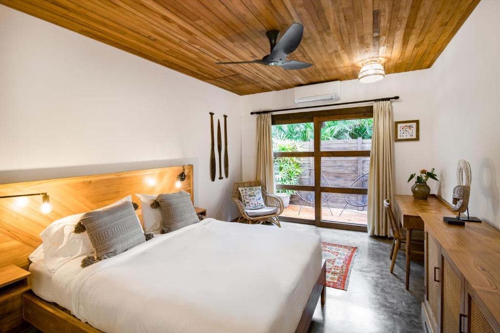 A cozy bedroom with a wooden ceiling and furniture, a large bed with white bedding and gray pillows, a ceiling fan, a desk, a chair, and glass doors leading to a lush outdoor garden area.