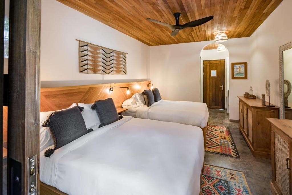 A cozy hotel room with two large beds, wooden ceiling, ceiling fan, decorative wall art, and a wooden dresser. The room has warm lighting, modern decor, and colorful patterned rugs on a concrete floor.