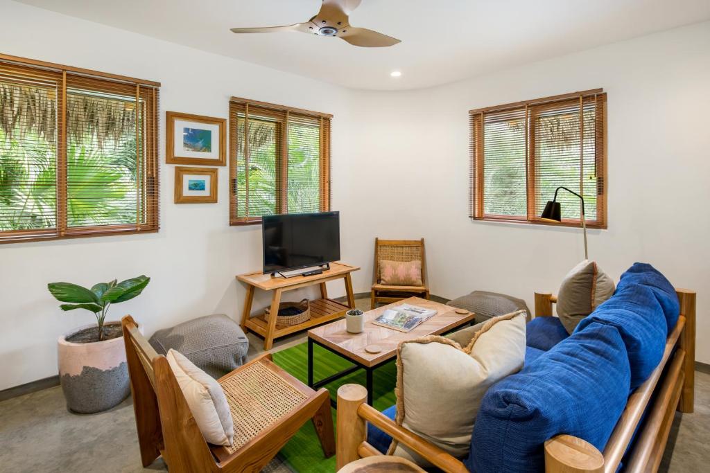 Cozy living room with wooden furniture, blue and beige cushions, a TV on a small stand, potted plant, and large windows with blinds letting in natural light. Green rug and artwork decorate the space.