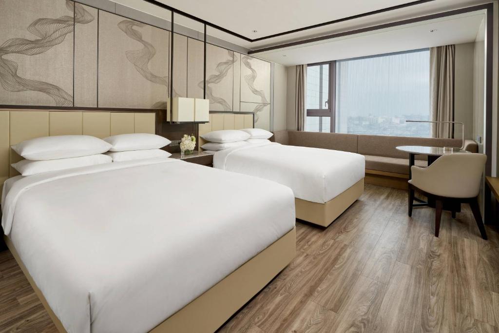 Modern hotel room with two large beds, a bench by a large window, a small round table with a chair, beige and wood decor, and abstract wall art behind the beds.