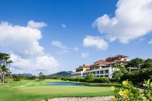 A large resort-style building with red-tiled roofs overlooks a golf course green and small pond, surrounded by lush greenery and set against a backdrop of hills under a bright blue sky with scattered clouds.