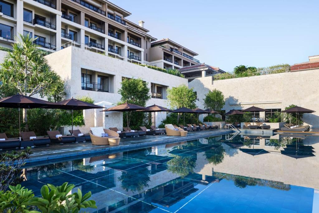 Outdoor swimming pool surrounded by lounge chairs and umbrellas, set in front of a modern, multi-story hotel with balconies and trees, under a clear blue sky.