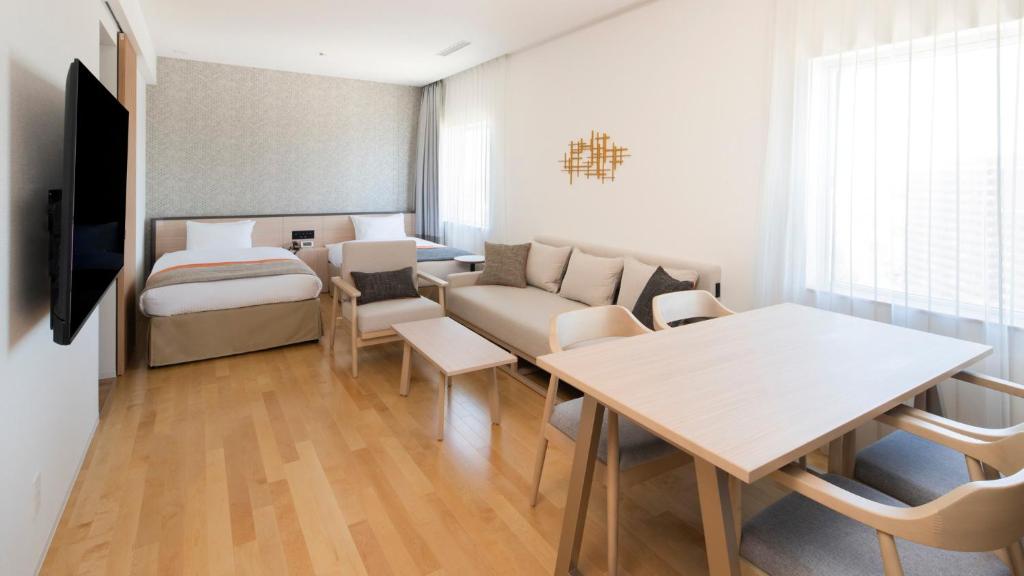 A modern studio apartment with light wood flooring, a bed, sofa, coffee table, dining table with four chairs, large window with sheer curtains, and minimalist decor.