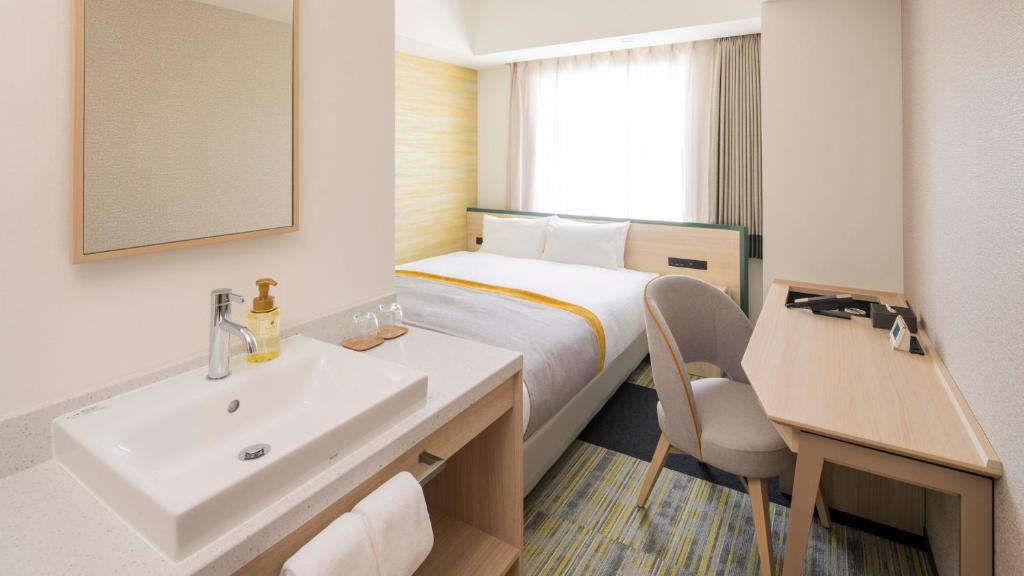 A modern, compact hotel room with a neatly made double bed, a wooden desk and chair, a wall-mounted TV, and a white sink with toiletries on the counter near the entrance. Natural light enters through a large window.