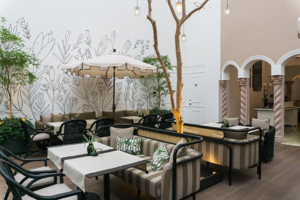 A stylish indoor cafe with striped seating, black chairs, potted plants, a central tree, and a large umbrella. The white wall features a minimalist botanical mural, and hanging lights add a cozy touch.