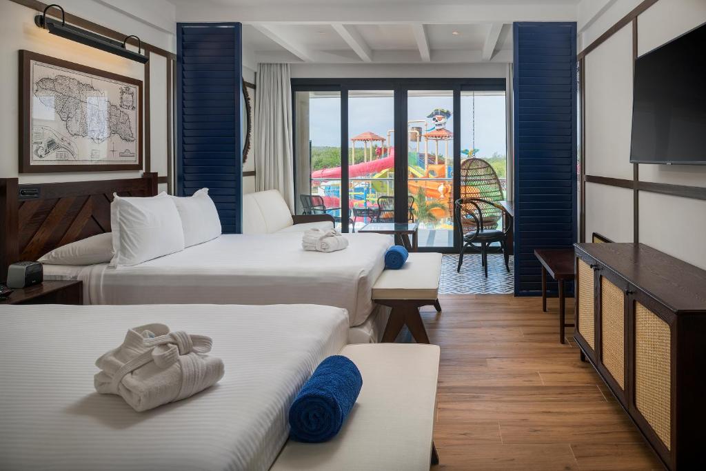 Modern hotel room with two double beds, white linens, blue accents, and a balcony view overlooking a colorful outdoor water park. Towels are placed on each bed, and a large TV is mounted on the wall.