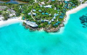 Aerial view of a tropical island with turquoise water, white sandy beaches, and green villas nestled among lush palm trees and vegetation along the coastline.