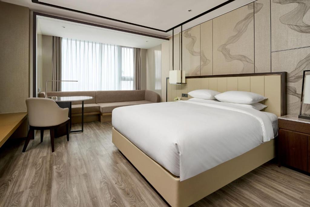 A modern hotel room with a large bed, white bedding, a wooden floor, a work desk with a chair, a built-in sofa by a window with sheer curtains, and neutral-toned walls with abstract designs.