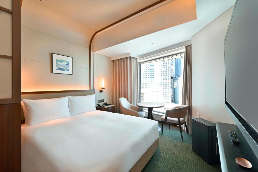 Modern hotel room with a large bed, a small round table and two chairs near a window overlooking city buildings, neutral-colored walls, and a wall-mounted TV.