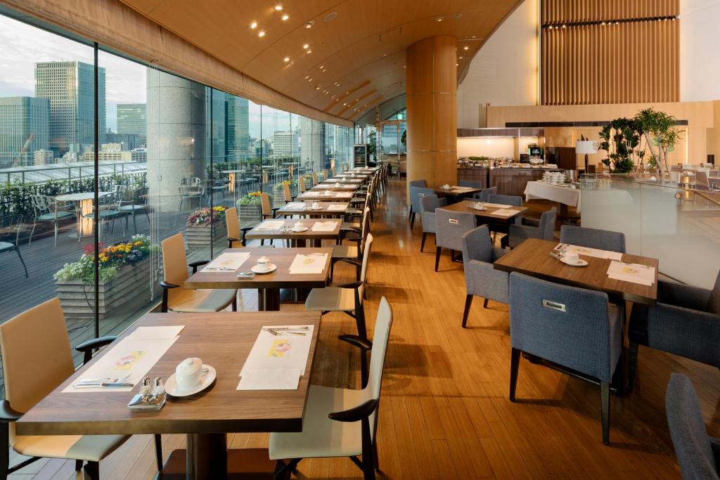 A modern, spacious restaurant with large windows offering a city view, wooden floors, neatly set tables with chairs, and bright natural light filling the room.