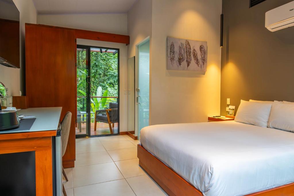 A modern hotel room with a large bed, white linens, wooden furniture, a small desk, and a balcony door open to greenery outside. Soft lighting creates a cozy and inviting atmosphere.