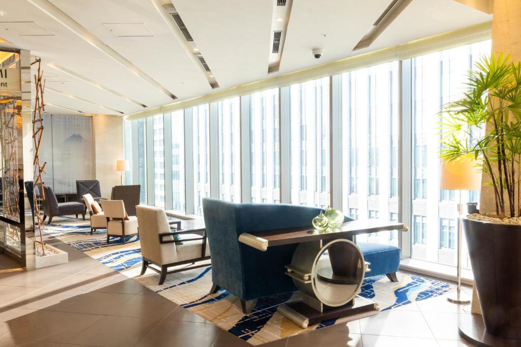 A modern lounge area with large floor-to-ceiling windows, blue and beige armchairs, a blue sofa, a contemporary table, decorative plants, and city buildings visible outside. Sunlight fills the bright, stylish room.