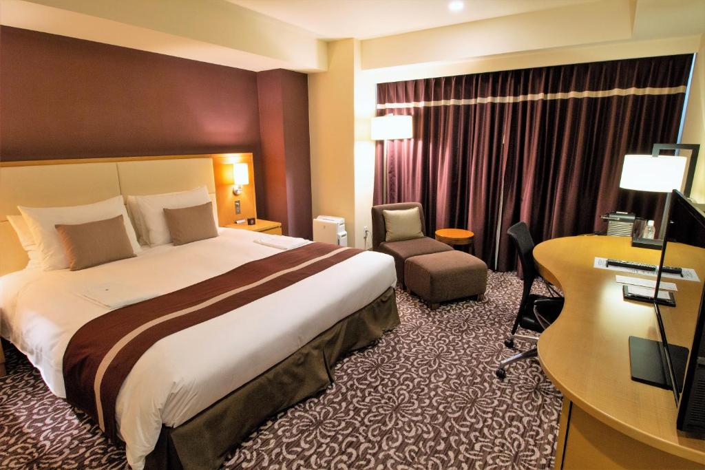 A modern hotel room with a large bed, two bedside lamps, an armchair with an ottoman, a desk with a chair, a flat-screen TV, patterned carpet, and dark curtains covering a window.