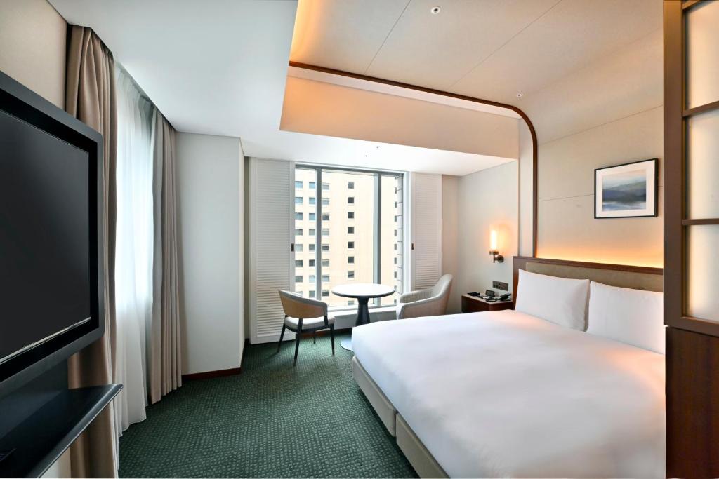 A modern hotel room with a large bed, a flat-screen TV, two chairs and a round table by a window, soft lighting, neutral decor, and a city view through the window.