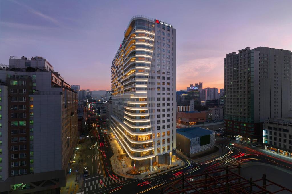 A tall, modern hotel building with Marriott signage is brightly lit at dusk in a busy city, surrounded by other high-rise buildings and streets with blurred car lights.