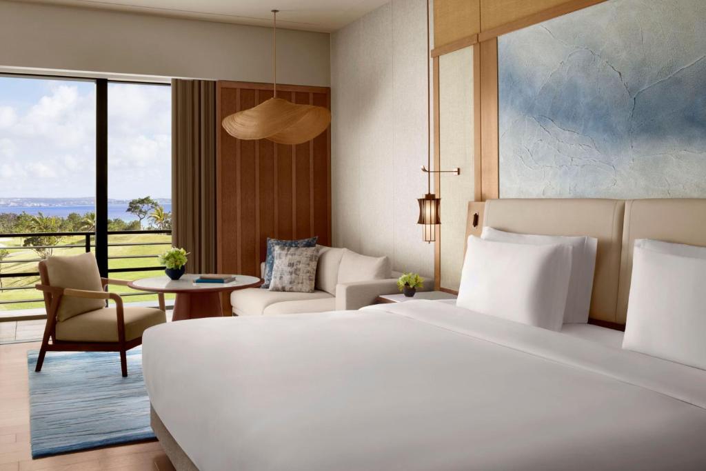 A modern hotel room with a large bed, white linens, a cozy seating area, a round table, floor-to-ceiling window with a scenic view of greenery and water, and stylish decor elements.