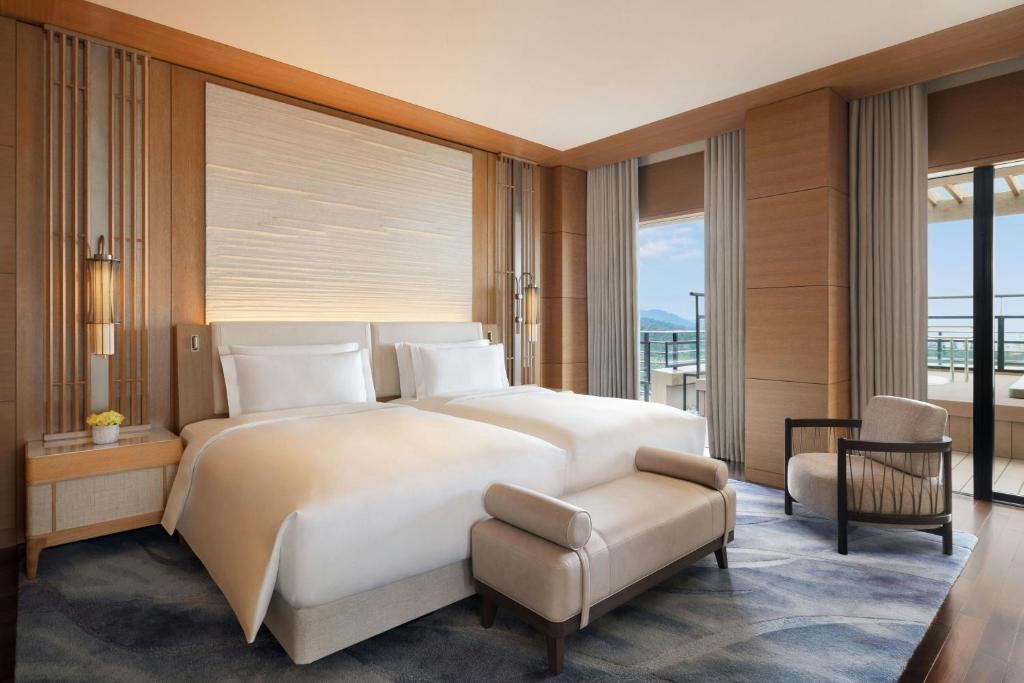A modern hotel room with a large bed, light wood paneling, soft lighting, a gray armchair, and glass doors opening to a balcony with views of the sea and distant hills.