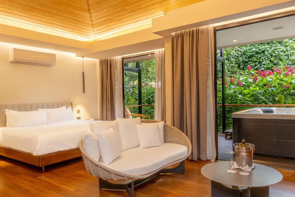 A modern hotel room with a double bed, a cozy white sofa, and a small round table with an ice bucket and glasses. Large sliding doors open to a patio with a hot tub and lush greenery outside.