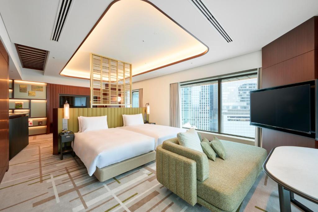 Modern hotel room with two double beds, a green sofa, large windows with city views, a wall-mounted TV, a small table, and wood-accented decor with warm lighting.